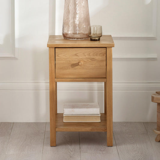Wooden side table with a drawer and a shelf, featuring a vase and a glass on top, against a white wall.
