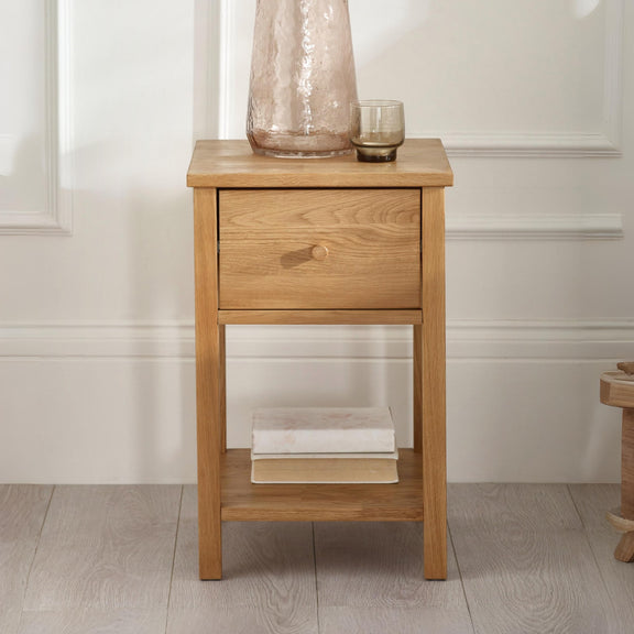 Wooden side table with a drawer and a shelf, featuring a vase and a glass on top, against a white wall.