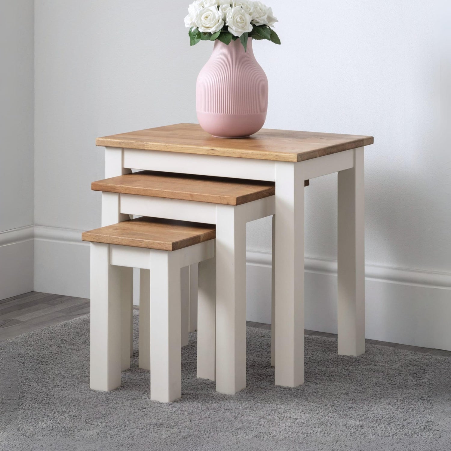 Set of three nesting tables with a pink vase and white flowers on top against a plain wall.