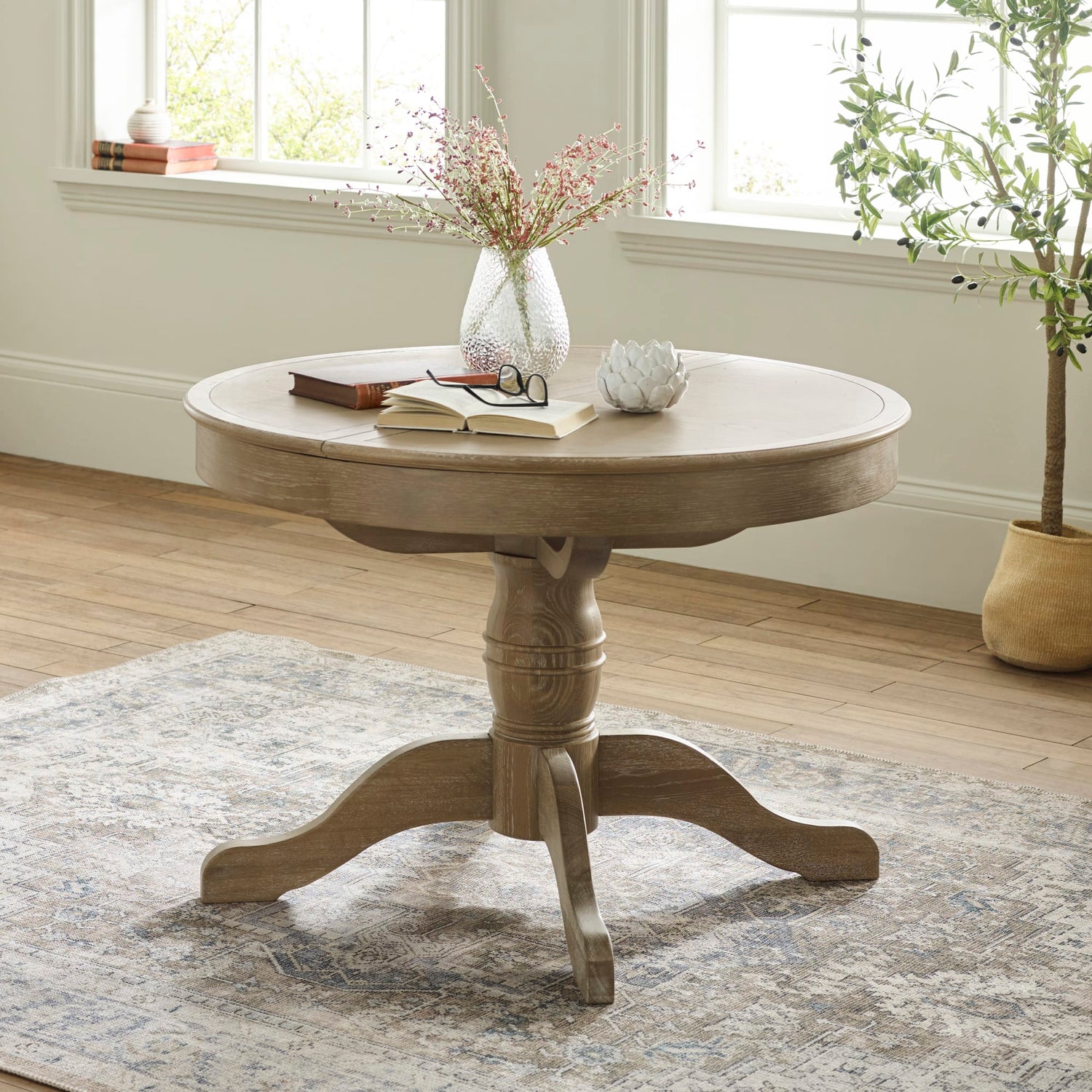 Round wooden table with decorative items in a room with windows and a plant.