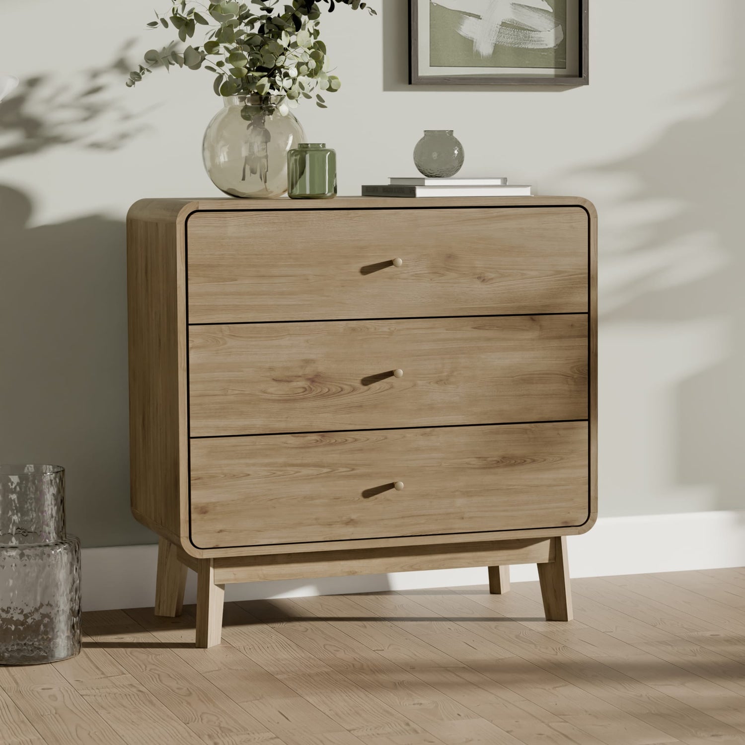 Wooden dresser with three drawers in a room with a vase of flowers and a framed picture on a shelf.