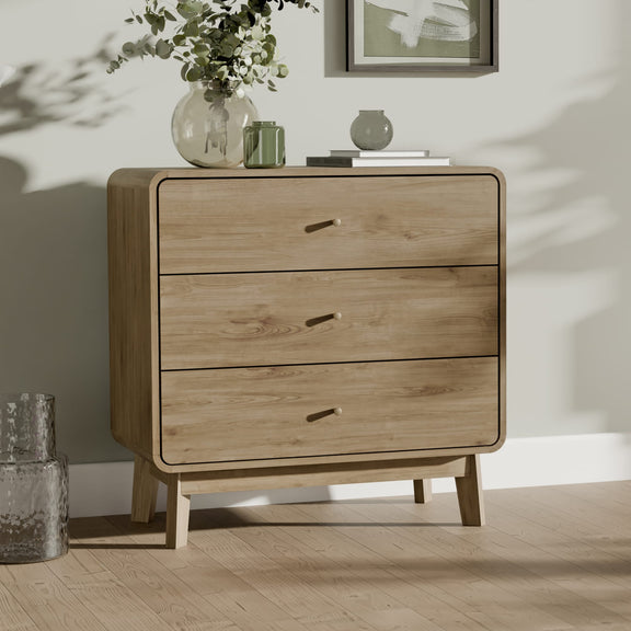 Wooden dresser with three drawers in a room with a vase of flowers and a framed picture on a shelf.