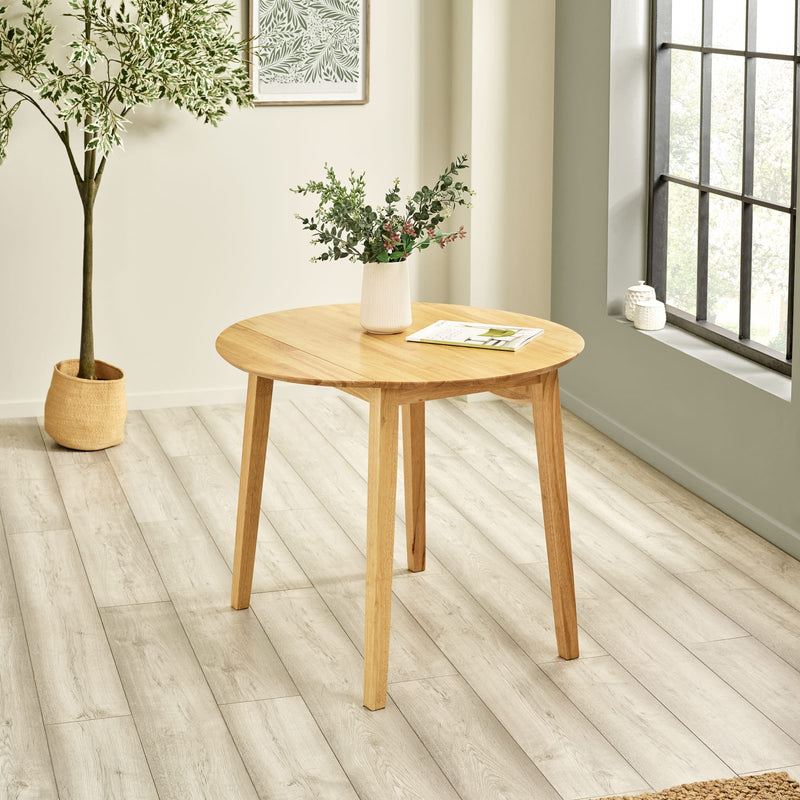 Wooden round table with plants in a room with light wood flooring and a window.