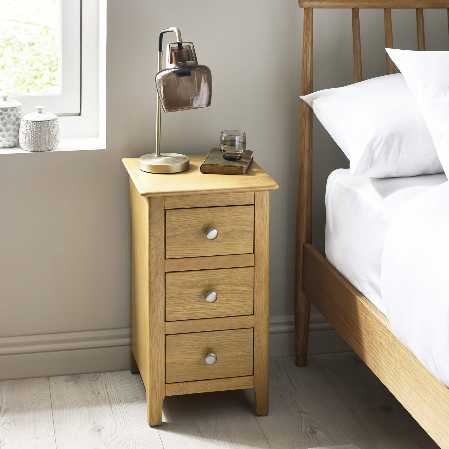 Wooden bedside table with lamp and drawers next to a bed in a bedroom.