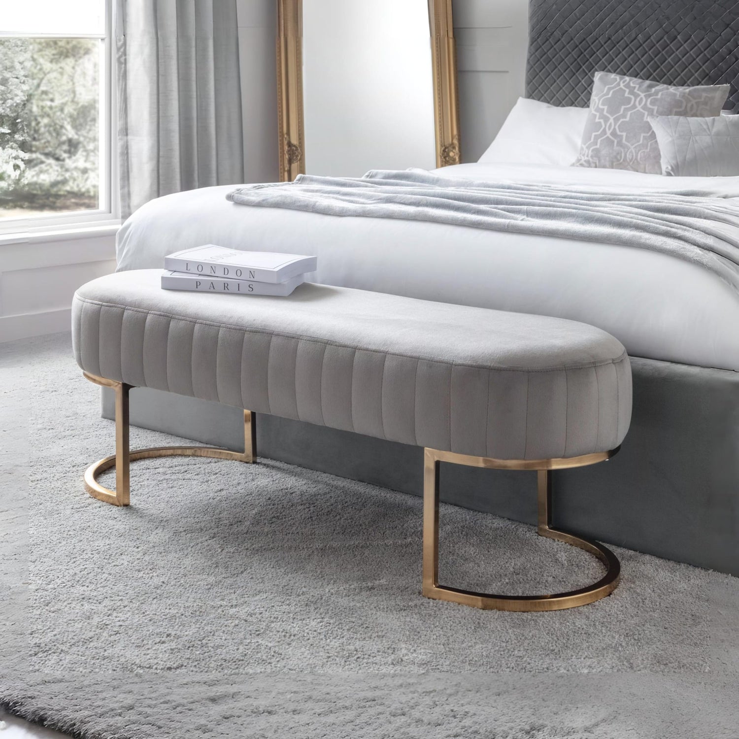 Gray upholstered bench with gold legs in front of a bed in a bedroom.