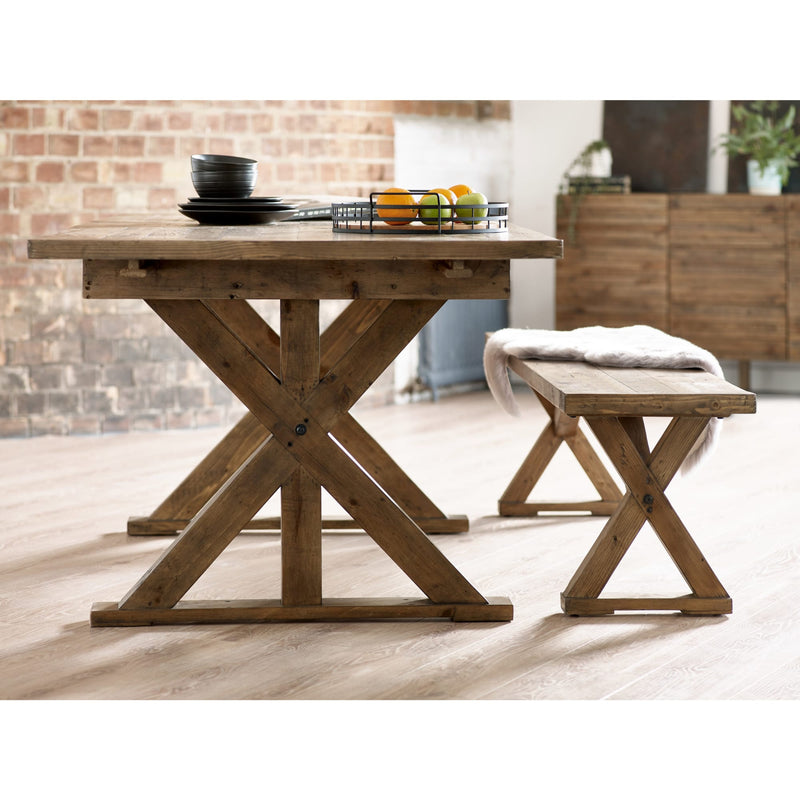Wooden dining table with X-shaped legs in a room with a brick wall and wooden bench.