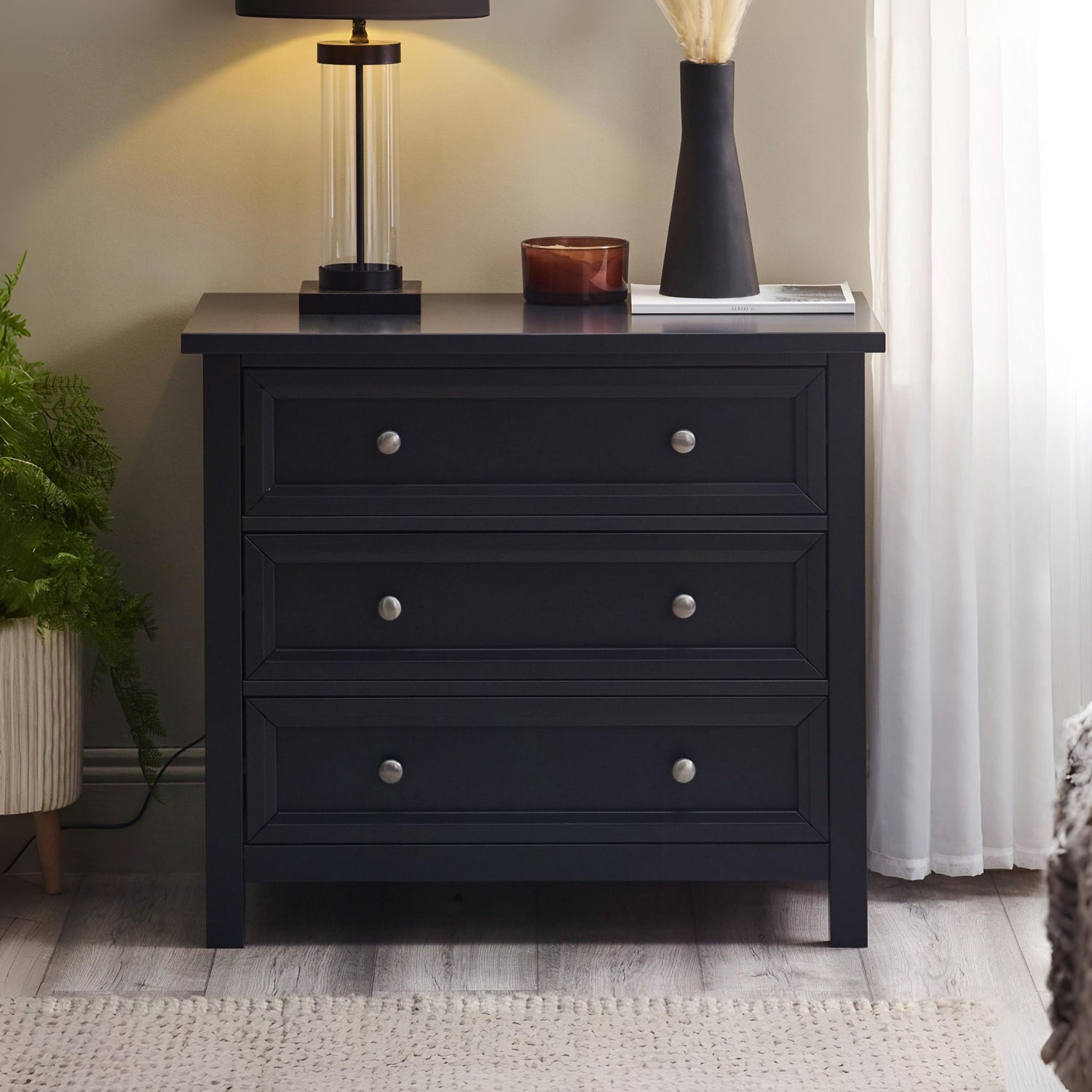 Black dresser with three drawers in a room setting with a lamp and plant.