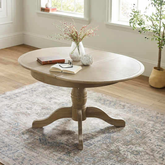 Round wooden table with books and a vase on a patterned rug in a room with windows and plants.