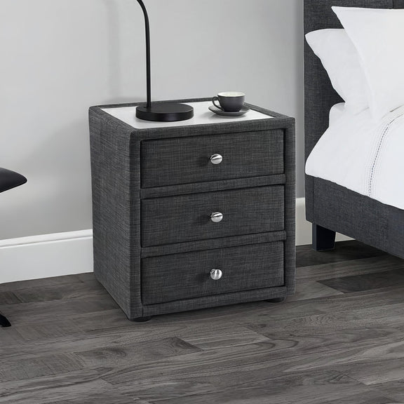 Gray textured nightstand with three drawers next to a bed in a bedroom setting.