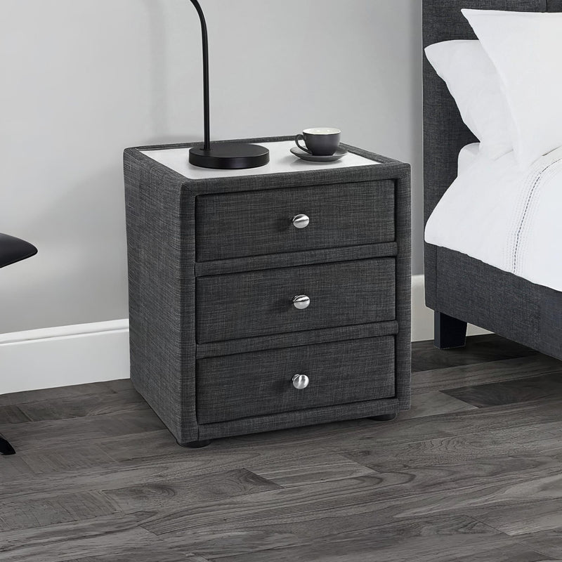Gray textured nightstand with three drawers next to a bed in a bedroom setting.