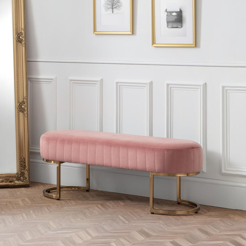 Pink velvet bench with gold legs in a room with white paneled walls and framed pictures.