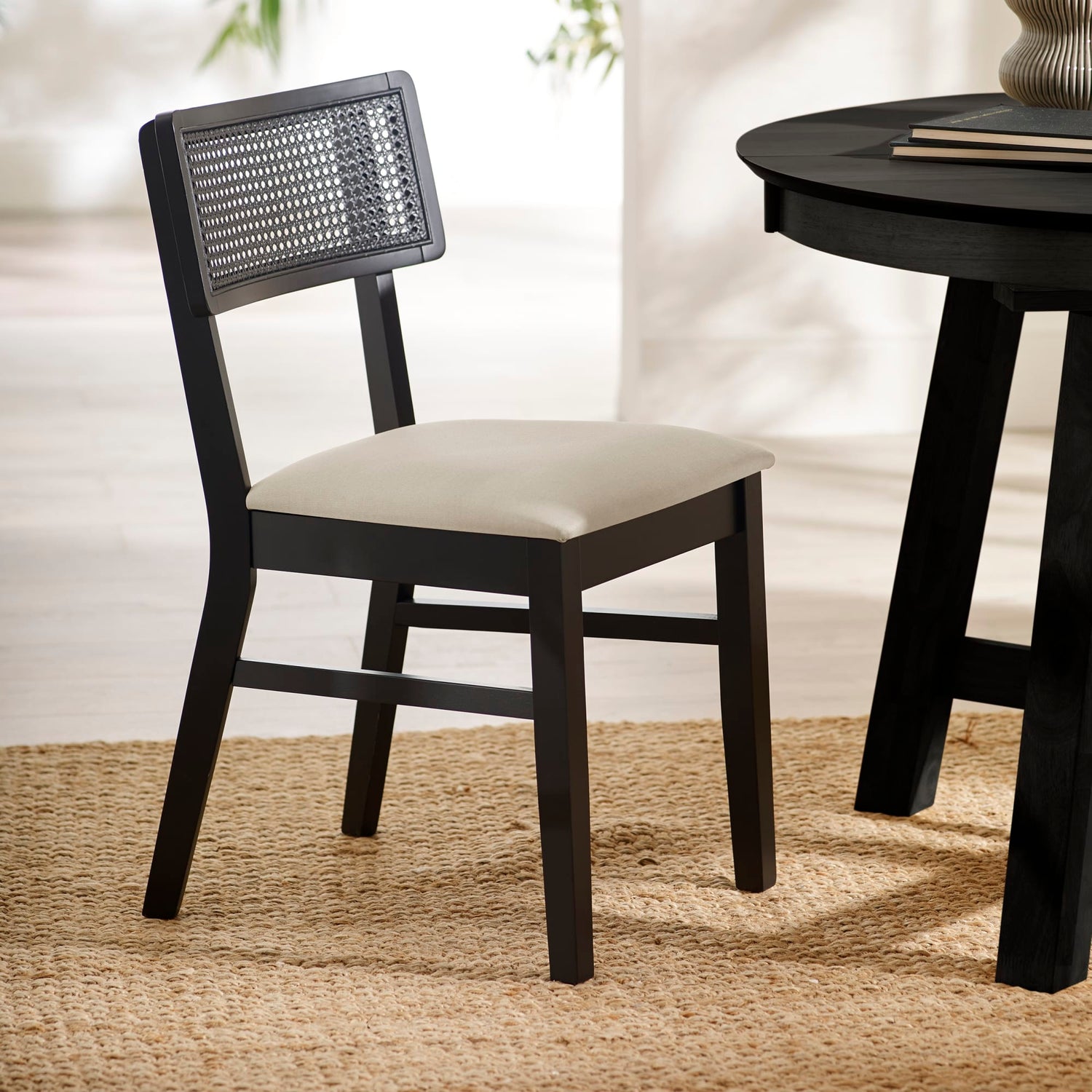 Black chair with woven backrest and white cushion next to a black round table on a beige rug.