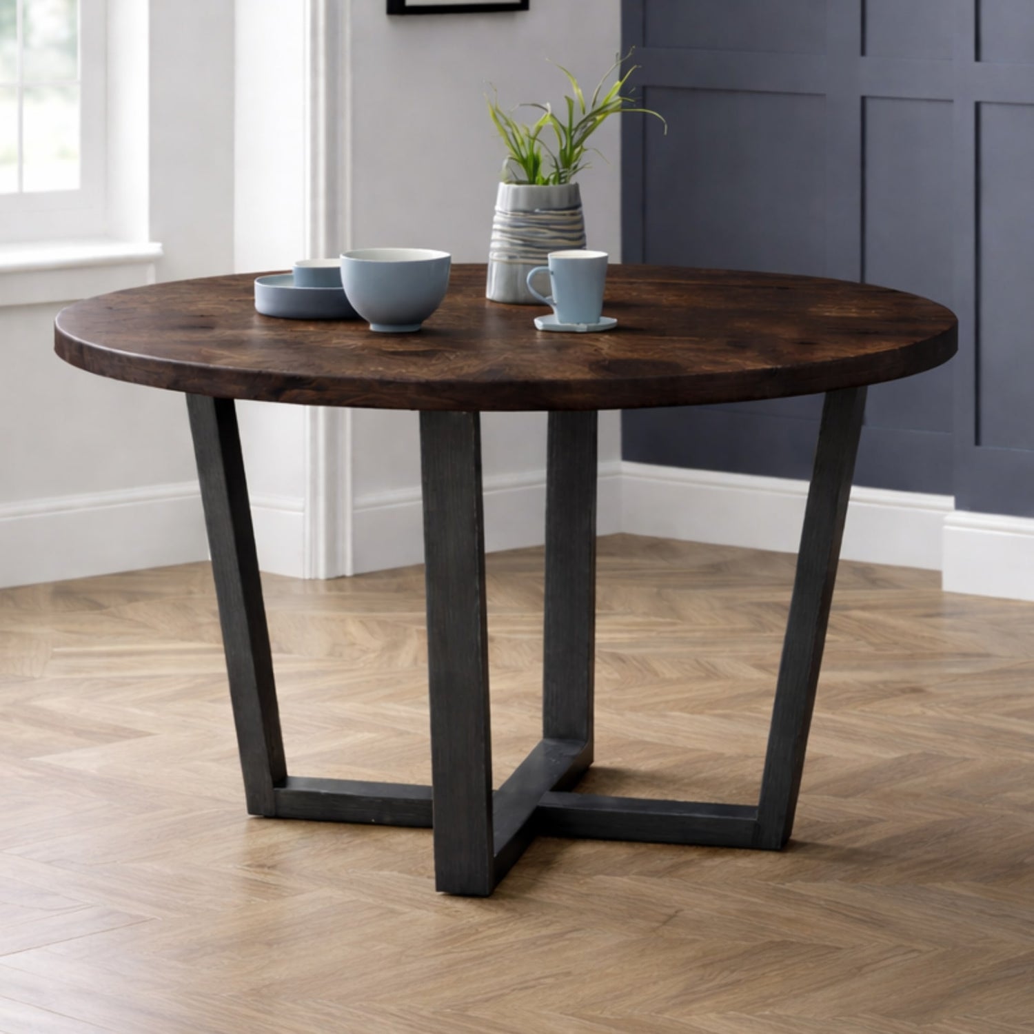 Round wooden table with dark metal legs in a room with a window and wall.