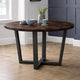 Round wooden table with dark metal legs in a room with a window and wall.
