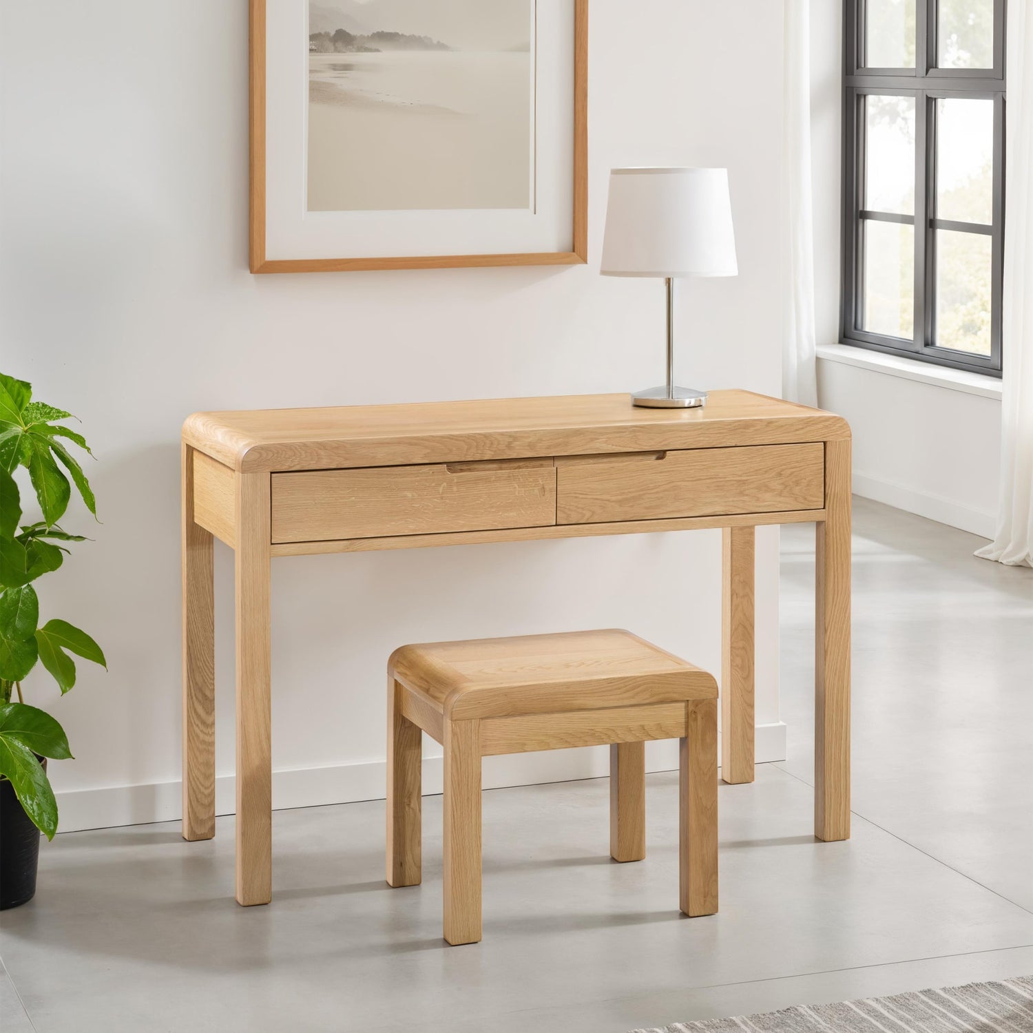 Wooden desk with two drawers and a matching stool in a room with a plant and framed picture.