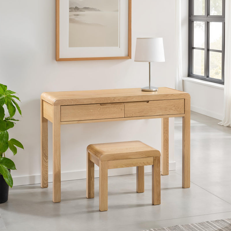 Wooden desk with two drawers and a matching stool in a room with a plant and framed picture.