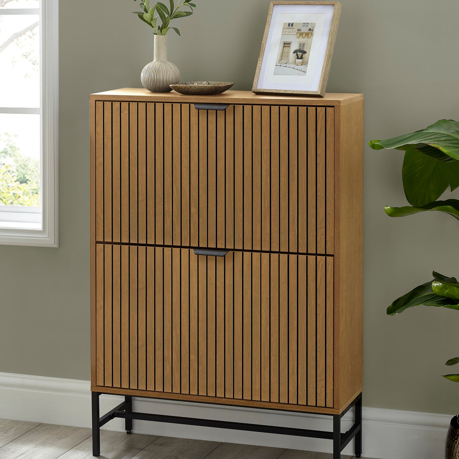 Wooden cabinet with two doors against a gray wall with a plant and framed picture on top.