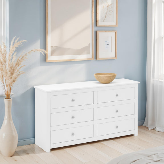 White dresser with six drawers in a room with light blue walls, framed artwork, and a vase with dried plants.