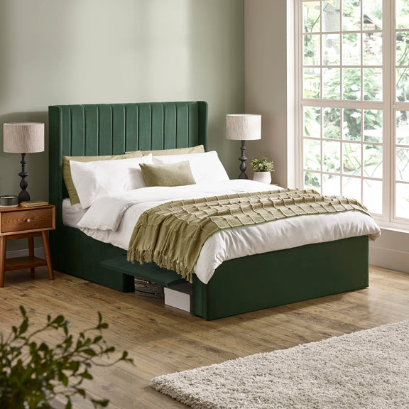 Green upholstered bed in a bedroom with wooden flooring and large windows.