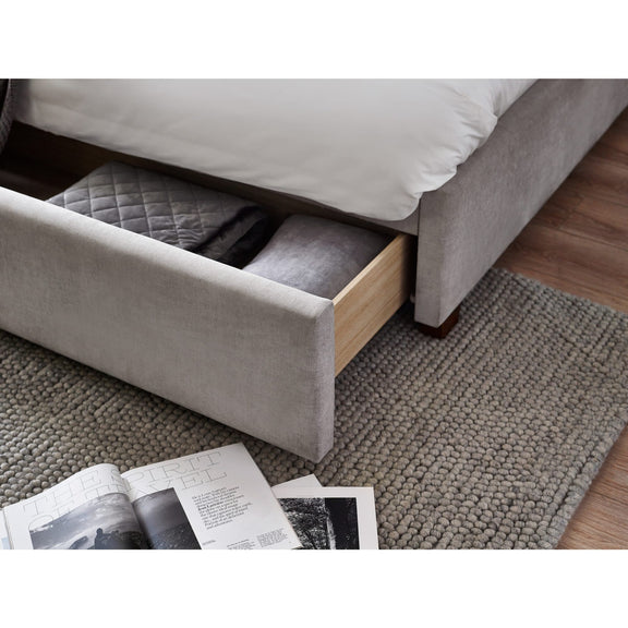 Gray upholstered bed with an open drawer revealing bedding on a carpeted floor.
