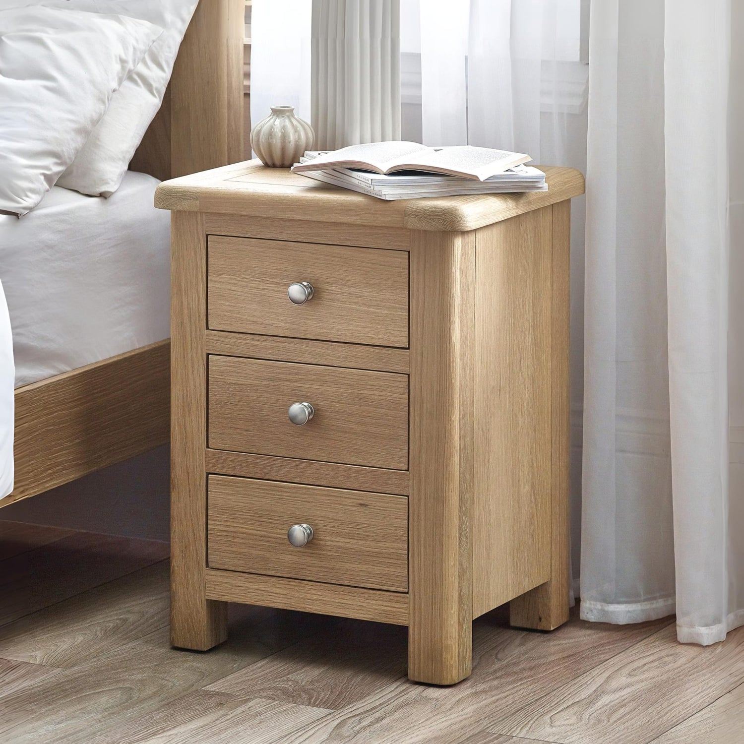 Wooden nightstand with three drawers next to a bed in a bedroom setting.