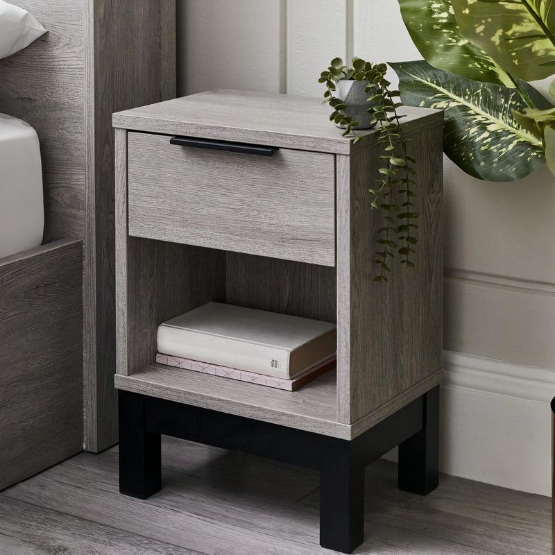 Wooden nightstand with a drawer and shelf, books, and a plant in a bedroom setting.