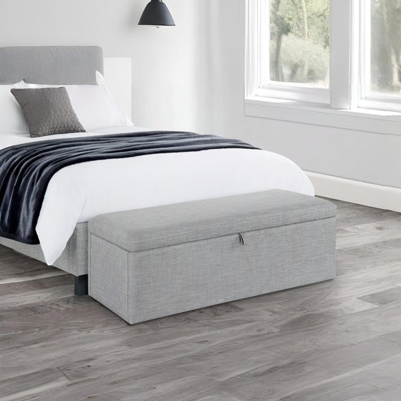 Gray upholstered storage bench in front of a bed with white bedding and gray blanket.