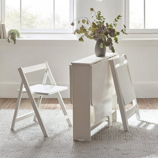 White folding table with two chairs in a bright room with large windows.