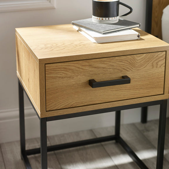 Wooden side table with a drawer and black metal legs, featuring a cup and books on top.