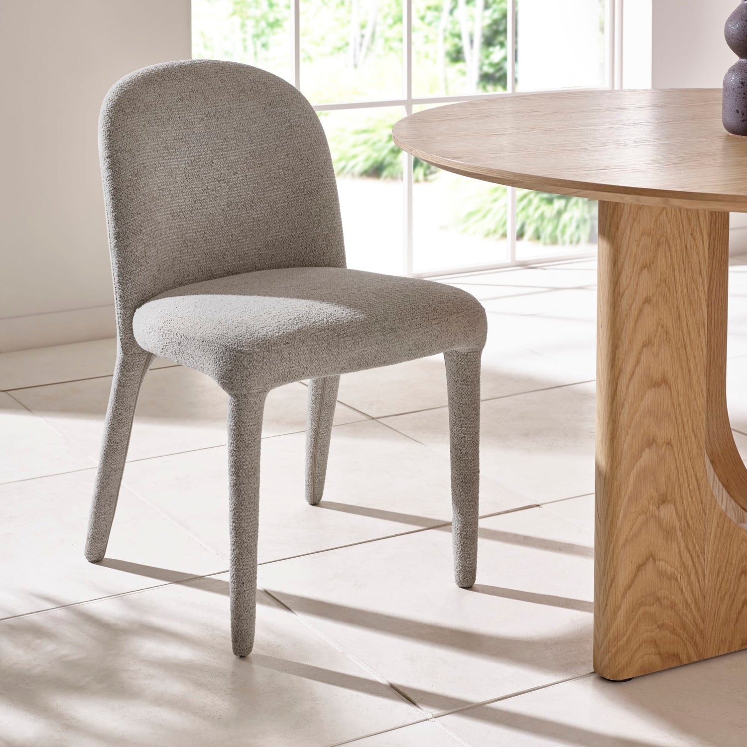 Gray upholstered chair next to a wooden table in a bright room with large windows.