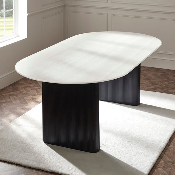 Modern dining table with black base and white marble top in a room with wooden flooring and a window.