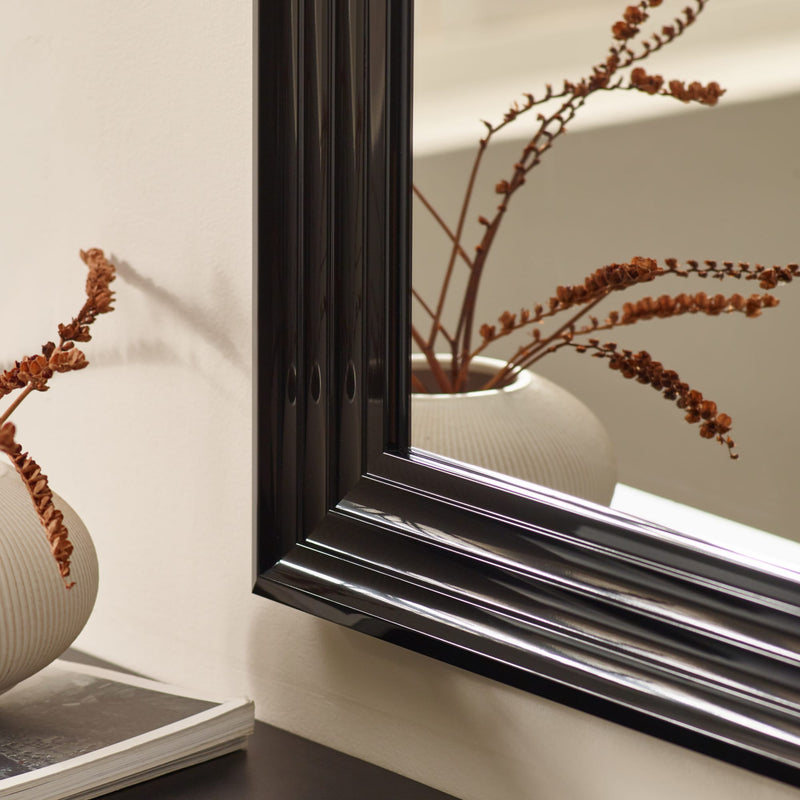 Decorative vase with branches inside a black-framed mirror on a beige wall.