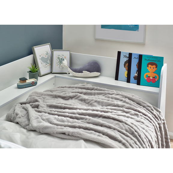 Gray blanket on a bed with a shelf above holding decorative items and books.