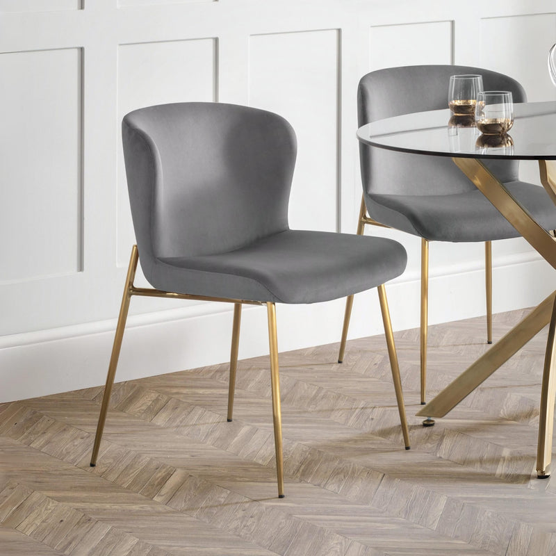 Gray chairs with gold legs around a glass dining table in a room with white paneled walls and wooden flooring.