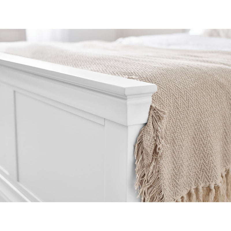 Beige throw blanket with fringe draped over a white bed frame.