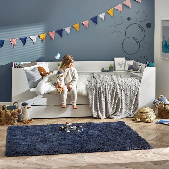 Child sitting on a bed in a bedroom with a blue rug and colorful flags on the wall.