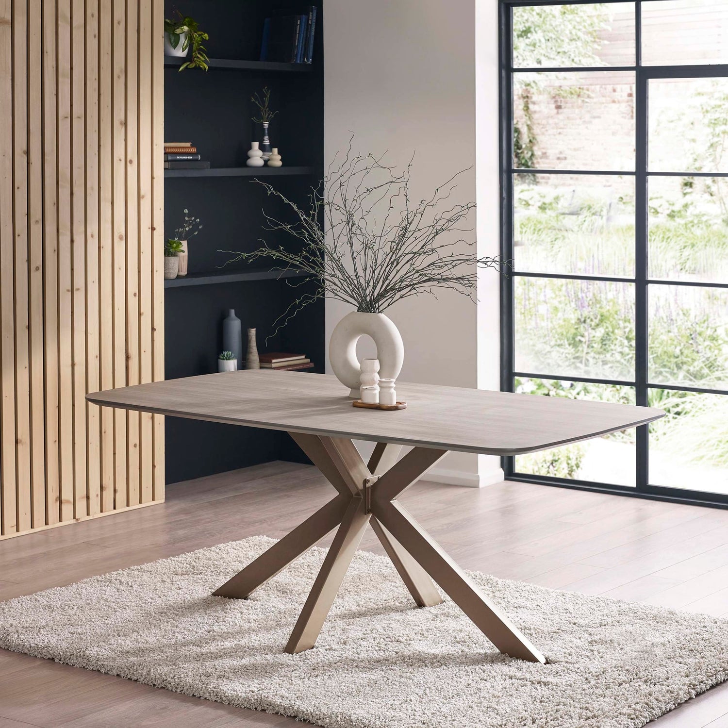 Modern dining table with wooden legs in a room with large windows and shelves.