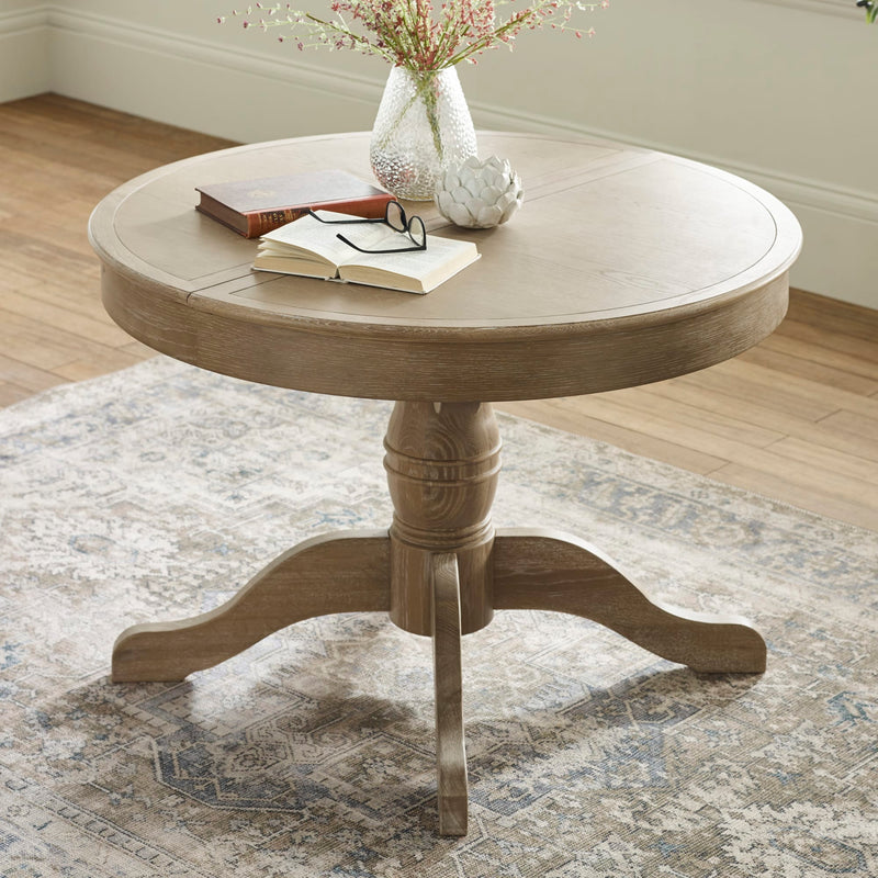 Round wooden table with a vase, book, and glasses on a patterned rug.