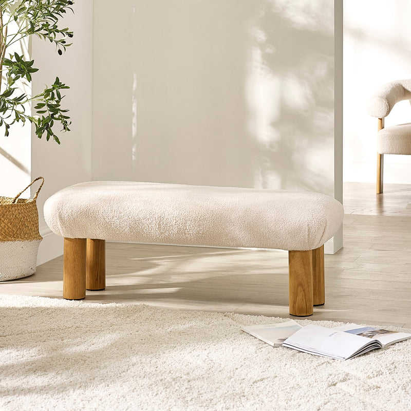 Beige ottoman with wooden legs in a room with a plant and magazine on the floor.
