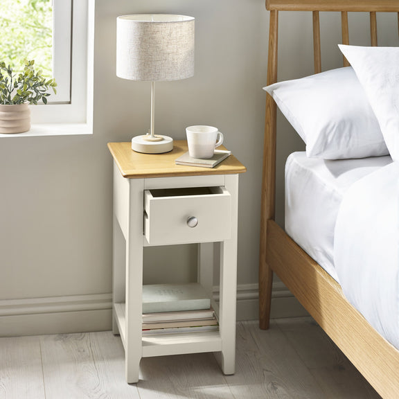 Compact white bedside cabinet with oak effect top placed next to a wooden bed