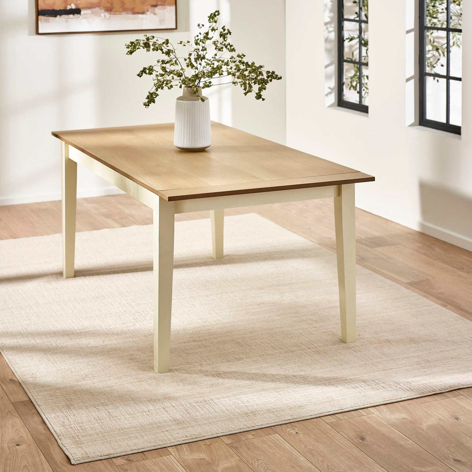 Wooden table with a vase of greenery on a light-colored rug in a room with a window.
