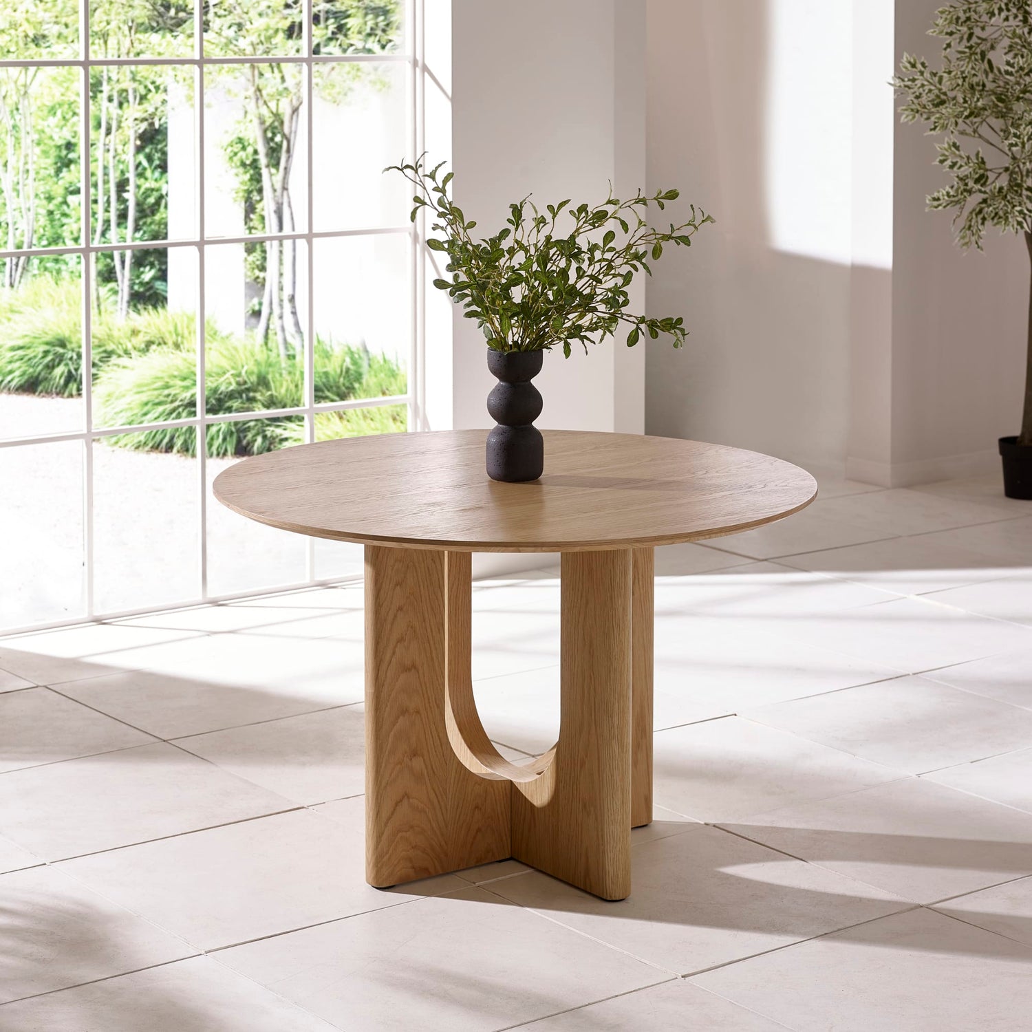 Wooden dining table with a vase of greenery in a bright room with large windows.