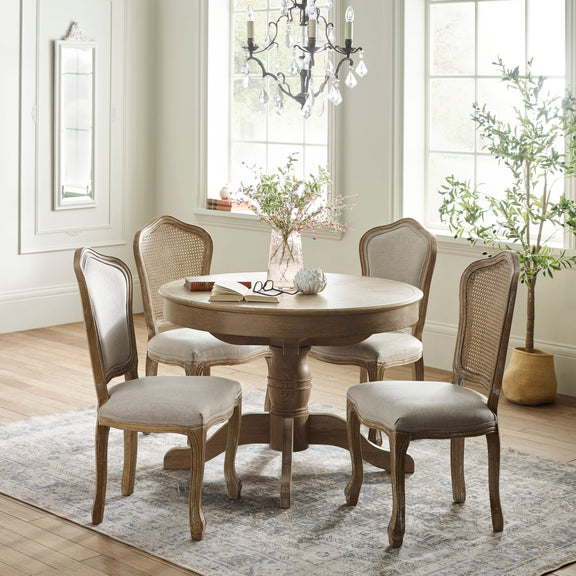 Round wooden dining table with four chairs in a well-lit room with large windows.