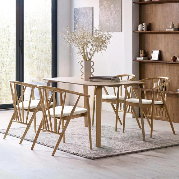 Dining room with wooden table and chairs on a rug, large windows, and shelves.