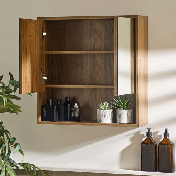 Wooden medicine cabinet with open door displaying bottles and plants, on a neutral wall.