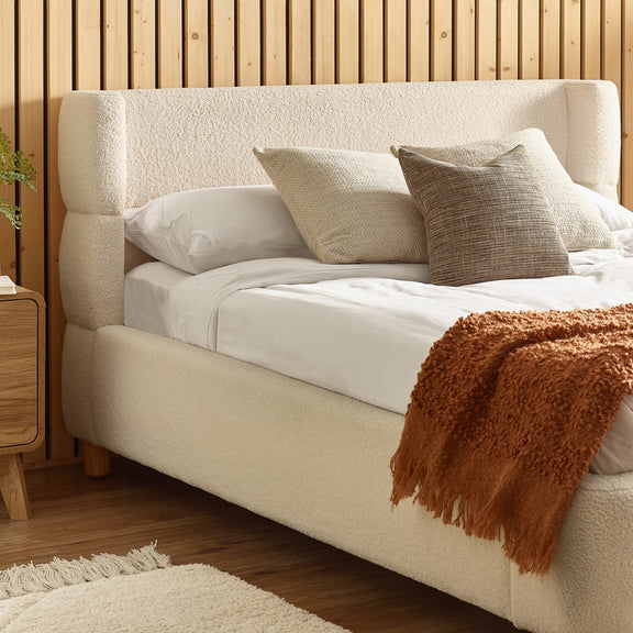 Beige upholstered bed with white bedding and brown throw blanket against a wooden wall.