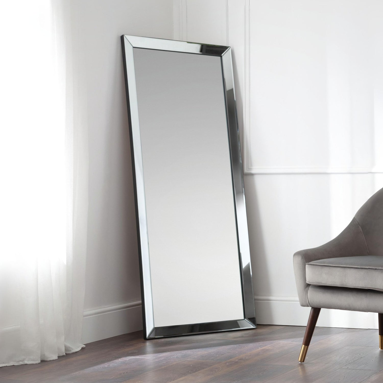 Large floor mirror with a modern frame leaning against a white wall in a room with a gray chair.