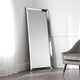 Large floor mirror with a modern frame leaning against a white wall in a room with a gray chair.