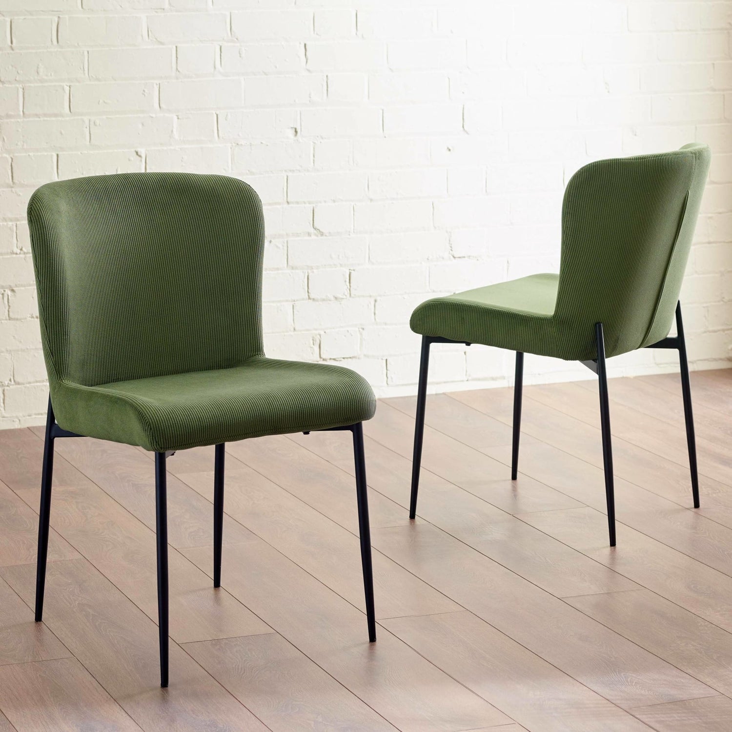 Two green chairs with black legs on a wooden floor against a white brick wall.