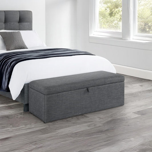 Gray storage bench in front of a bed in a bedroom with wooden flooring.